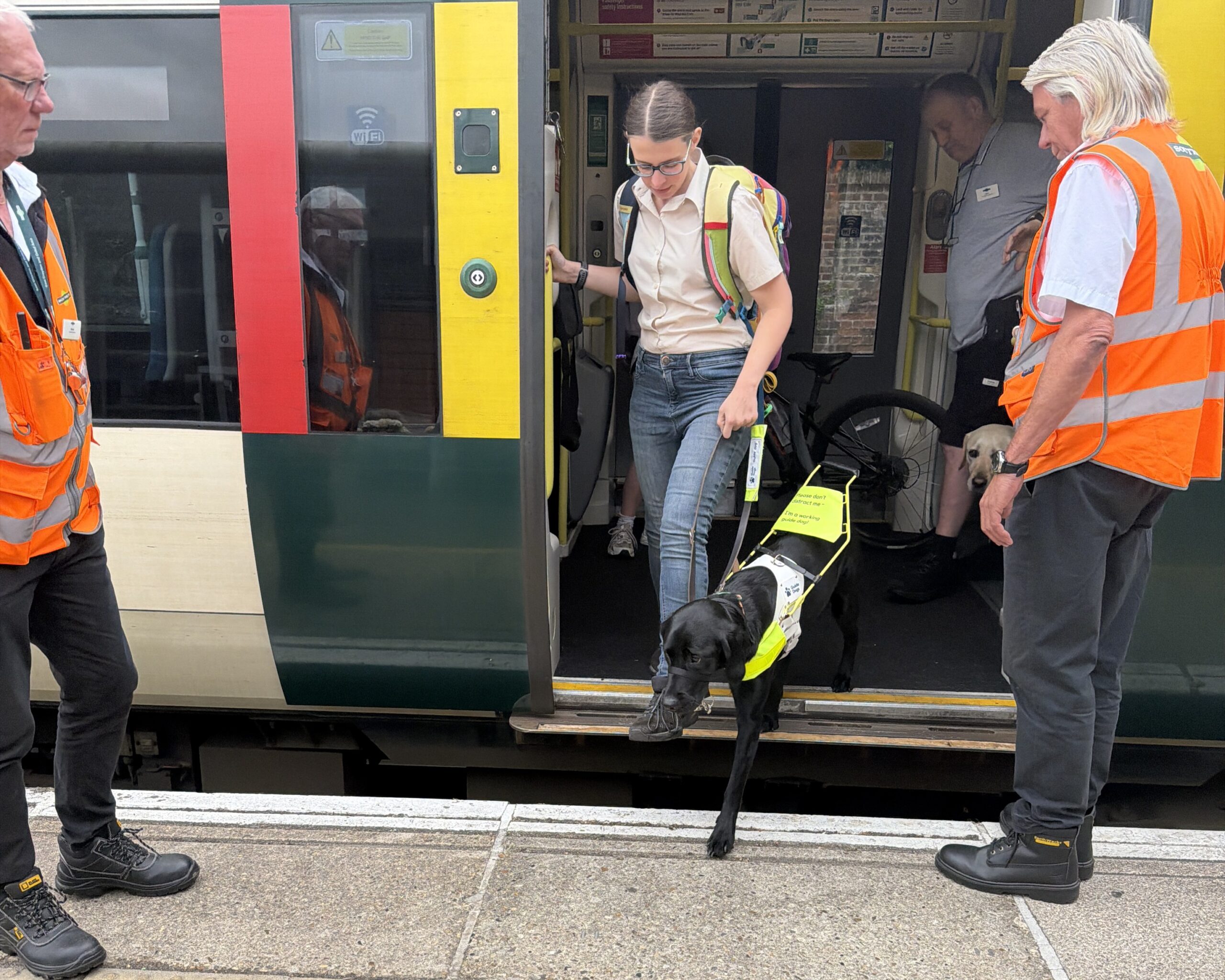 Lauren, SLC Coordinator for South East England, steps off a Southern train with her guide dog, as part of the GTR project. Lauren holds onto the dog's harness as they navigate the gap between the train and the platform. Two staff members, wearing orange hi-visibility jackets stand on either side of the train doorway.