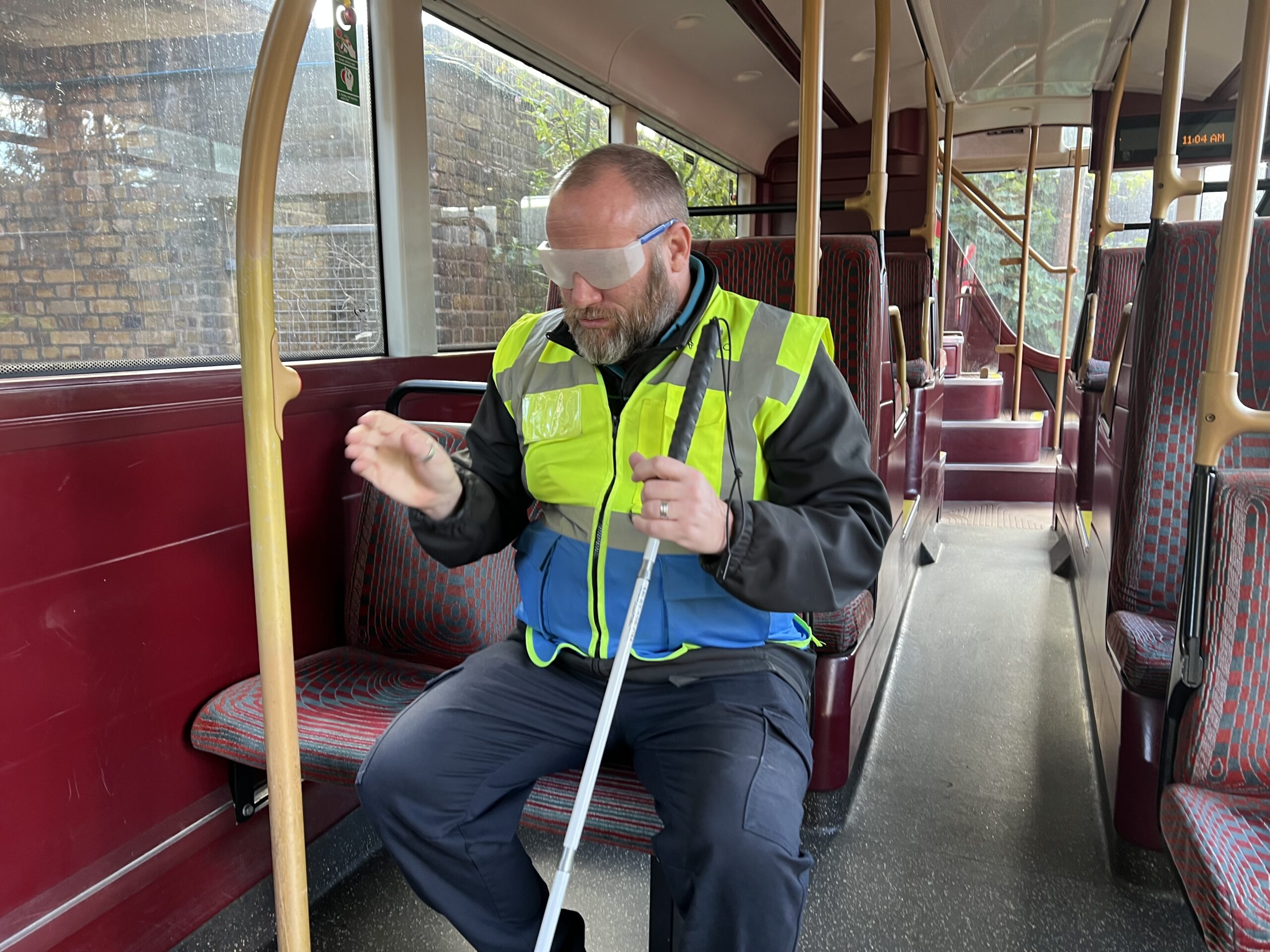 Noel, a bus driver at Arriva's Clapton Bus Garage, sits on a stationary bus wearing sight loss simulation glasses and holding a long mobility cane. Reaching forward to locate the stop bell, Noel takes part in an exercise designed to simulate the experience of a blind or partially sighted passenger travelling independently by bus.
