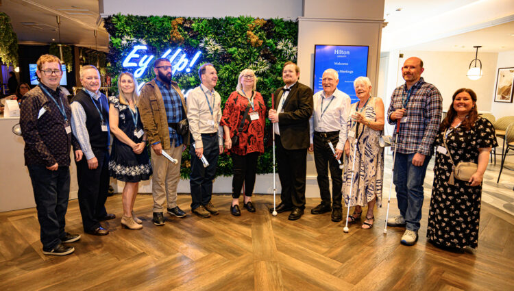 Group shot of Bedfordshire and Hertfordshire SLC taken at the 2026 Sight Loss Councils Conference. SLC members stand in a semi-circle in front of a neon light which reads 'Ey Up!"