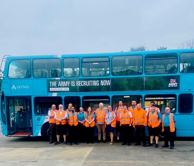 A group photo taken outside a blue Arriva double-decker bus at Arriva Yorkshire's Dewsbury depot. Sight Loss Council volunteers, Guide Dogs staff. and Arriva employees stand together in front of the bus. All wear high-visibility orange vests after taking part in a bus driver vision awareness training session aimed at improving accessibility for blind and partially sighted passengers.
