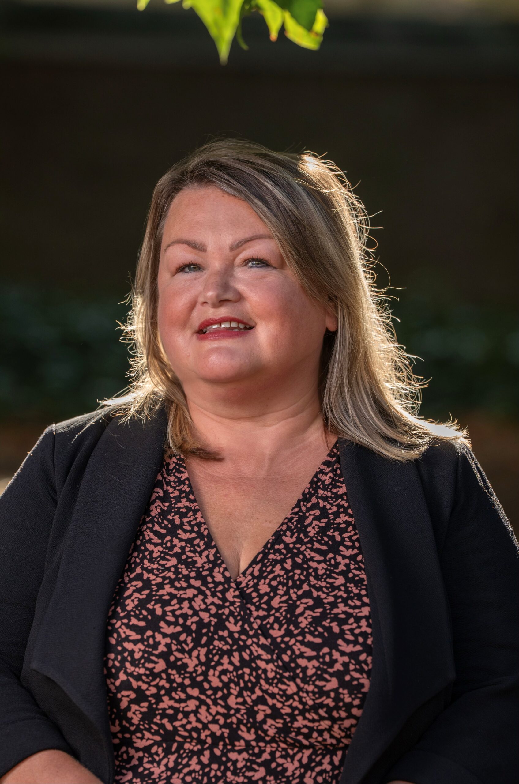 An outdoors portrait style photo of Lydia Horbory, CEO of Bus Users UK. She has shoulder-length light-coloured hair, wearing a black blazer, and a black-and-pink patterned dress. She is seated in warm sunlight against a softly blurred background of greenery and fencing.
