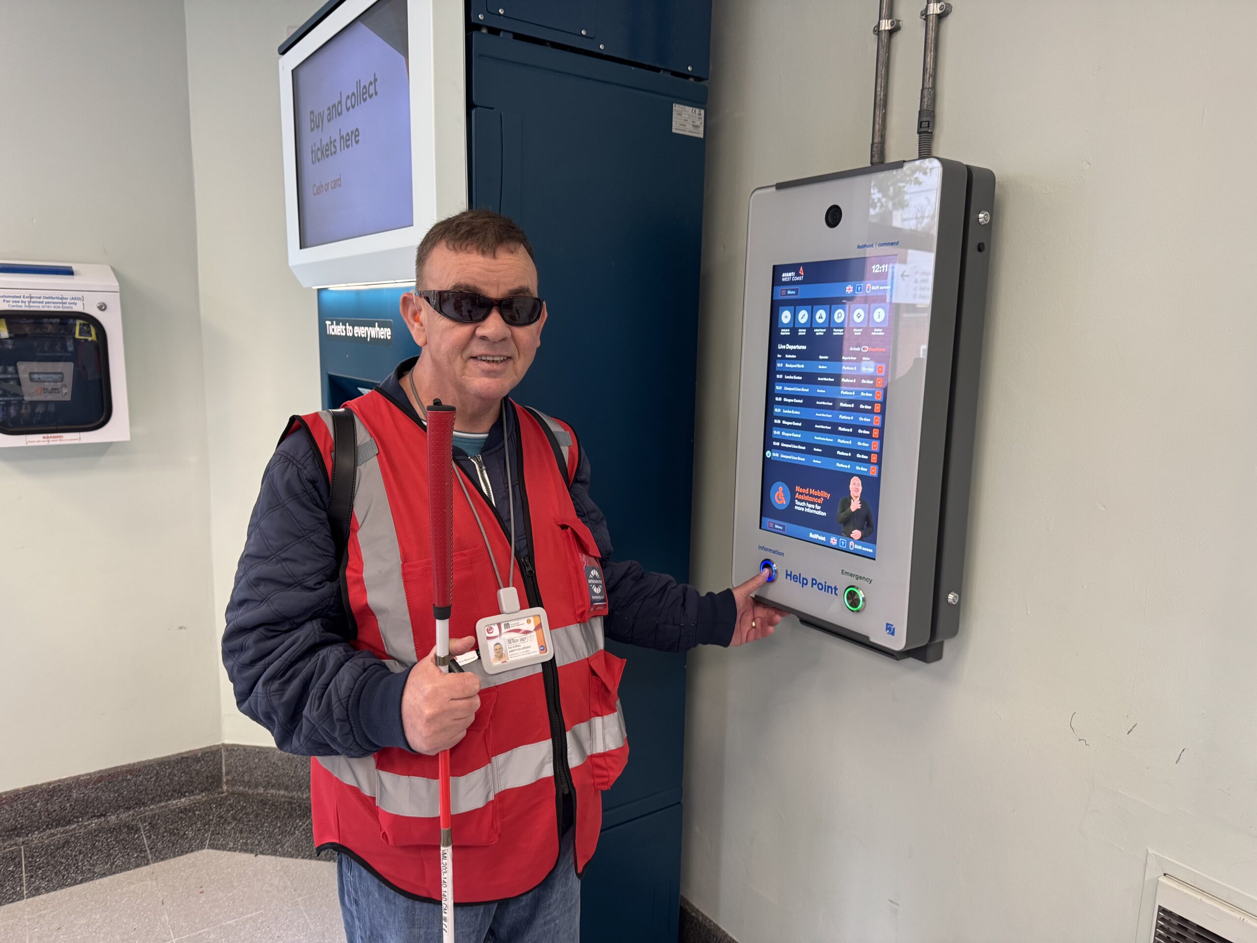 A SLC volunteer, wearing a red high-visibility vest, dark glasses and an ID badge stands beside a touchscreen help point at a railway station. He is holding a long white cane and pressing a button on the help point. A ticket machine is visible in the background.