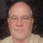 A head-and-shoulders portrait of Ian, Sussex Sight Loss Council member. He is wearing glasses and a light-coloured shirt, looking towards the camera. The photograph is taken indoors against a dark background.
