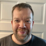 Head-and-shoulders portrait of Calum McCready, a Yorkshire and Humberside Sight Loss Council volunteer, standing indoors in front of a white panelled door and wearing a dark grey T-shirt.