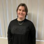 Klaudia, Staffordshire SLC member, stands indoors in front of white vertical blinds, wearing a black DKNY jumper with a subtle logo detail and a silver necklace. A grey radiator is visible to one side.