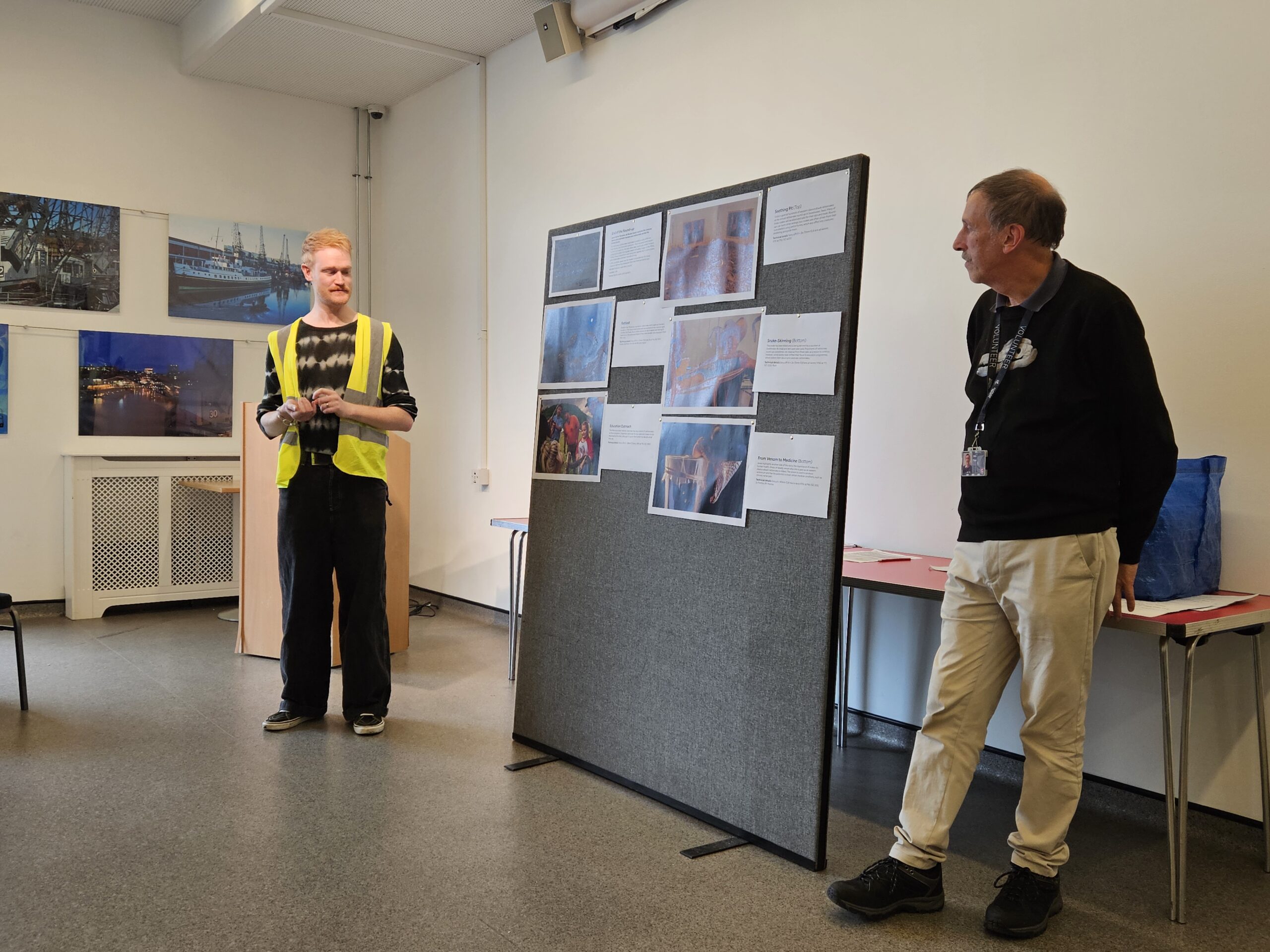 Two presenters stand beside a display board of exhibition images and accompanying descriptions during a live audio description session.