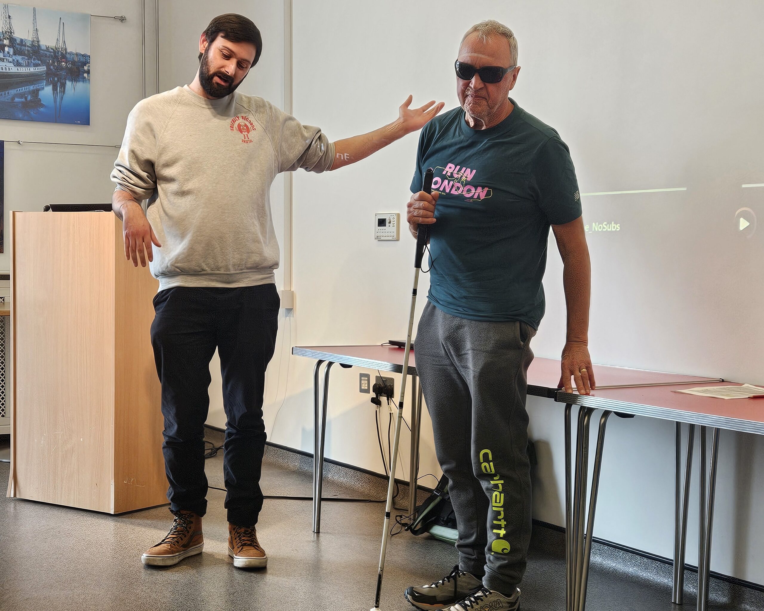 Phil Harrison stands holding a long white cane while filmmaker Reuben Armstrong speaks beside him during a discussion about their documentary, An Exercise in Perspective.