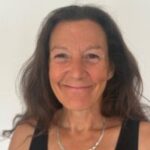 Head-and-shoulders portrait of Sacha, Sussex SLC member. She has long dark hair and is wearing a black sleeveless top and a silver chain necklace, standing against a plain light-coloured background and facing the camera. The image is well lit with a clear, neutral backdrop.