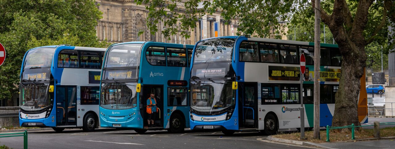 Three blue double-decker buses parked at a city-centre bus stop beneath large leafy trees. A historic stone building stands in the background, with bus destination displays and roadside signs visible. One bus has its doors open while a member of staff stands nearby.