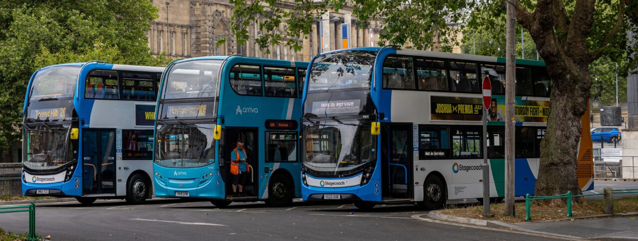 Three blue double-decker buses parked at a city-centre bus stop beneath large leafy trees. A historic stone building stands in the background, with bus destination displays and roadside signs visible. One bus has its doors open while a member of staff stands nearby.