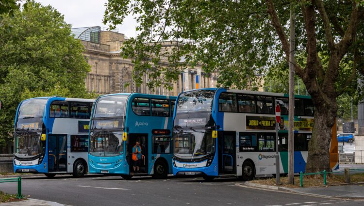 Three blue double-decker buses parked at a city-centre bus stop beneath large leafy trees. A historic stone building stands in the background, with bus destination displays and roadside signs visible. One bus has its doors open while a member of staff stands nearby.