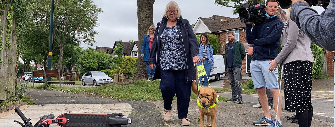 a guide dog user walks by a badly parked e-scooter, other people are standing by and watching.