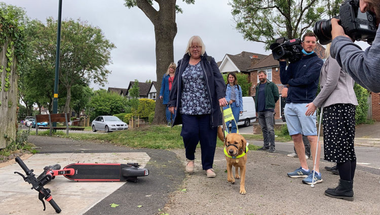 a guide dog user walks by a badly parked e-scooter, other people are standing by and watching.