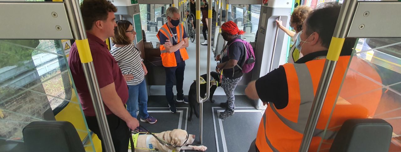 A group of people inside a train, some are guide dog users, some are wearing high-vis vests.