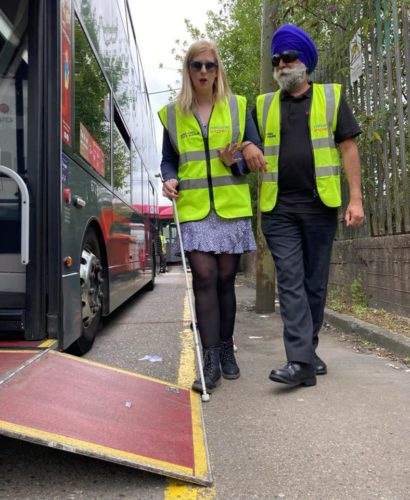 Two people wearing Hi-vis jackets. A cane user is guiding a man wearing sim-specs get onto a bus.