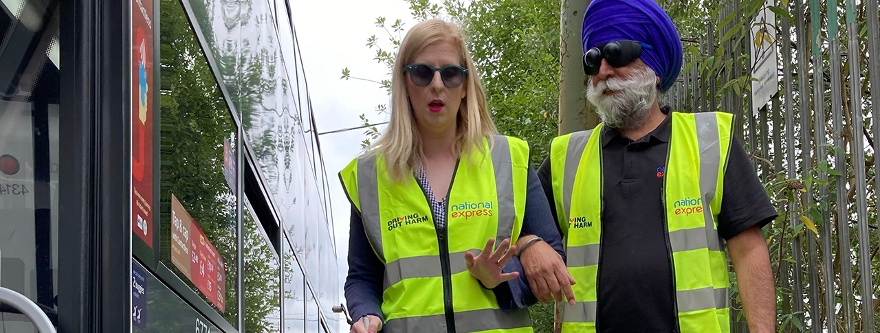 A woman with a white cane guiding a man wearing sim specs by a bus, both wearing high vis vests