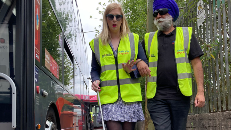 A woman with a white cane guiding a man wearing sim specs by a bus, both wearing high vis vests
