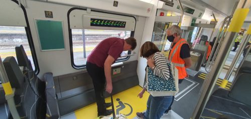 Two people examine the electric sockets in the wheelchair area of a train carriage, a man in a high-vis vest is standing by