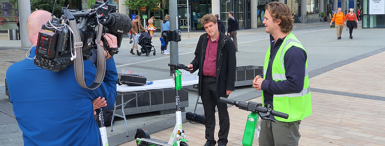 A group of people standing in a public square by several electric scooters. Among whom include a man in a high vis vest, a cameraman, and a reporter.