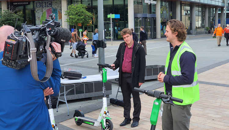 A group of people standing in a public square by several electric scooters. Among whom include a man in a high vis vest, a cameraman, and a reporter.