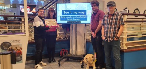 Greater Manchester Sight Loss Council members smiling and standing beside a screen that is showing a film titled 'See it my way' - Sight Loss Councils celebrate Purple Tuesday' Two members are also holding a giant cookie that reads 'Celebrating Purple Tuesday!' 