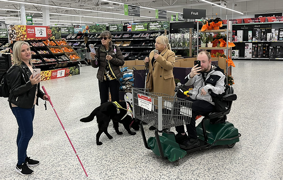 Engagement Manager Kelly Barton pictured with three Sight Loss Council members testing out the GoodMaps Explore app in an Asda store. Aisles of groceries are in the background.