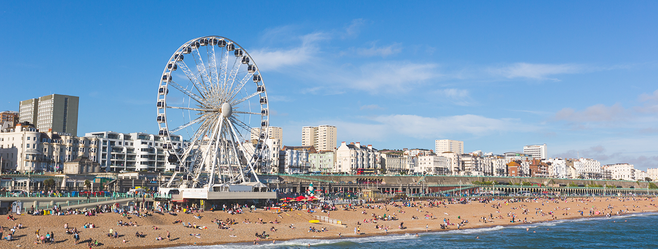 Brighton view of seaside from the pier. Panoramic shot with the famous ferris wheel, the stoney beach with unrecognisable persons on a sunny summer day.