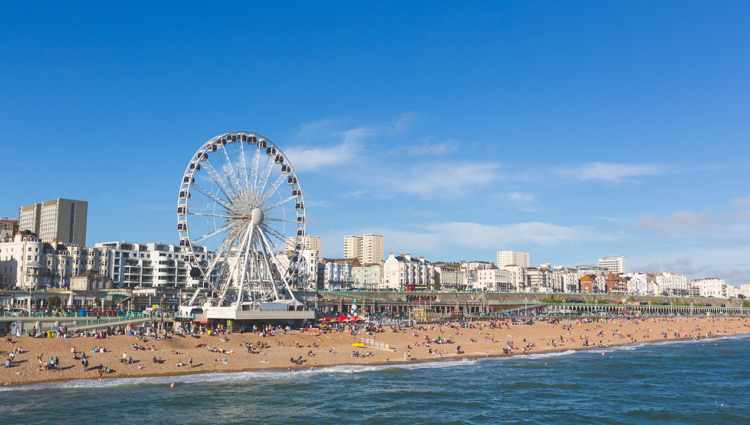 Brighton view of seaside from the pier. Panoramic shot with the famous ferris wheel, the stoney beach with unrecognisable persons on a sunny summer day.