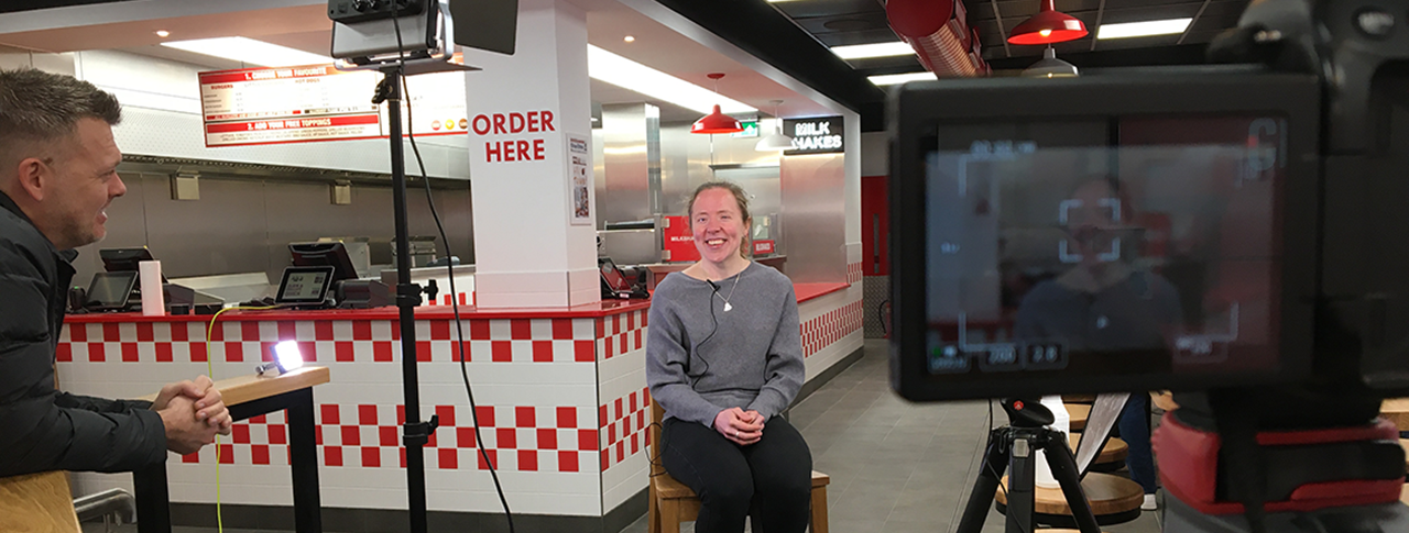 Lora smiling, sat on a stool in a Five Guys restaurant in front of camera equipment