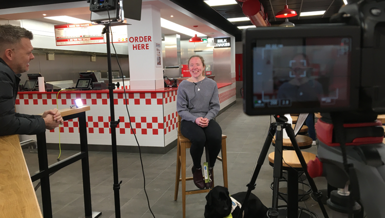 Lora smiling sat on a stool in a Five Guys restaurant in from of various filming equipment