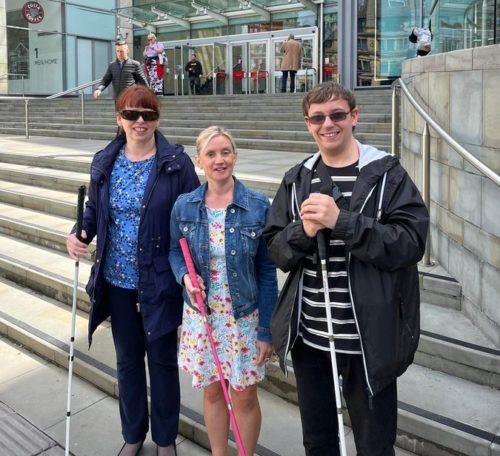 Kelly is stood holding her pink cane abs sight loss council members Anthony and Mary are either side of her and are at the bottom of the steps which lead up to the Arndale Centre.
