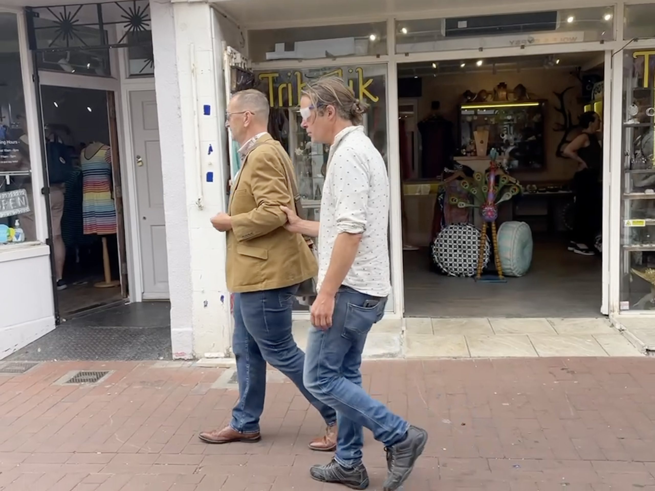 Cllr Jamie Lloyd being guided by Daniel Brookbanks, CEO of East Sussex Vision Support. Jamie is holding onto Daniel's elbow. He is wearing a white shirt and jeans, Daniel a brown blazer and jeans. Interiors shops in the background.
