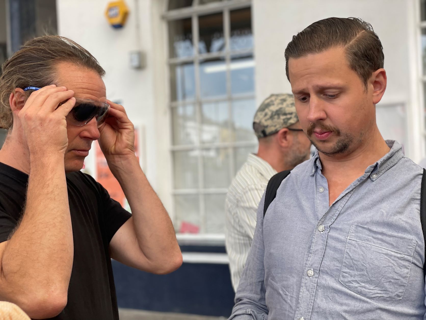 Close up of Councillor Jamie Lloyd outside Brighton train station, trying on a pair of simulation specs, standing with James Beckett, Dave Smith's support worker. James is wearing a blue shirt, looking down.