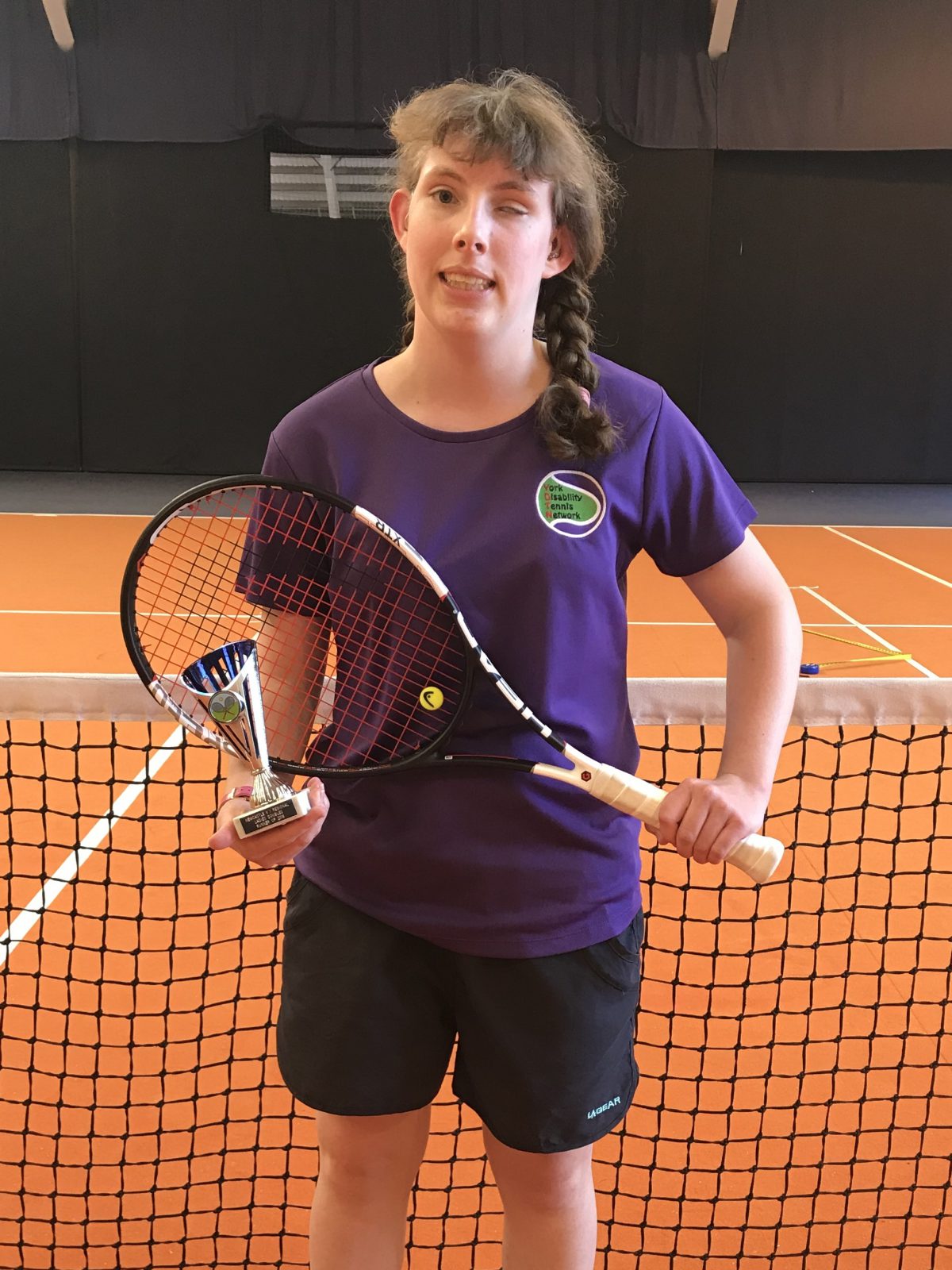 Lydia Wrightson holding a tennis racket in a tennis court, holding one of her awards.
