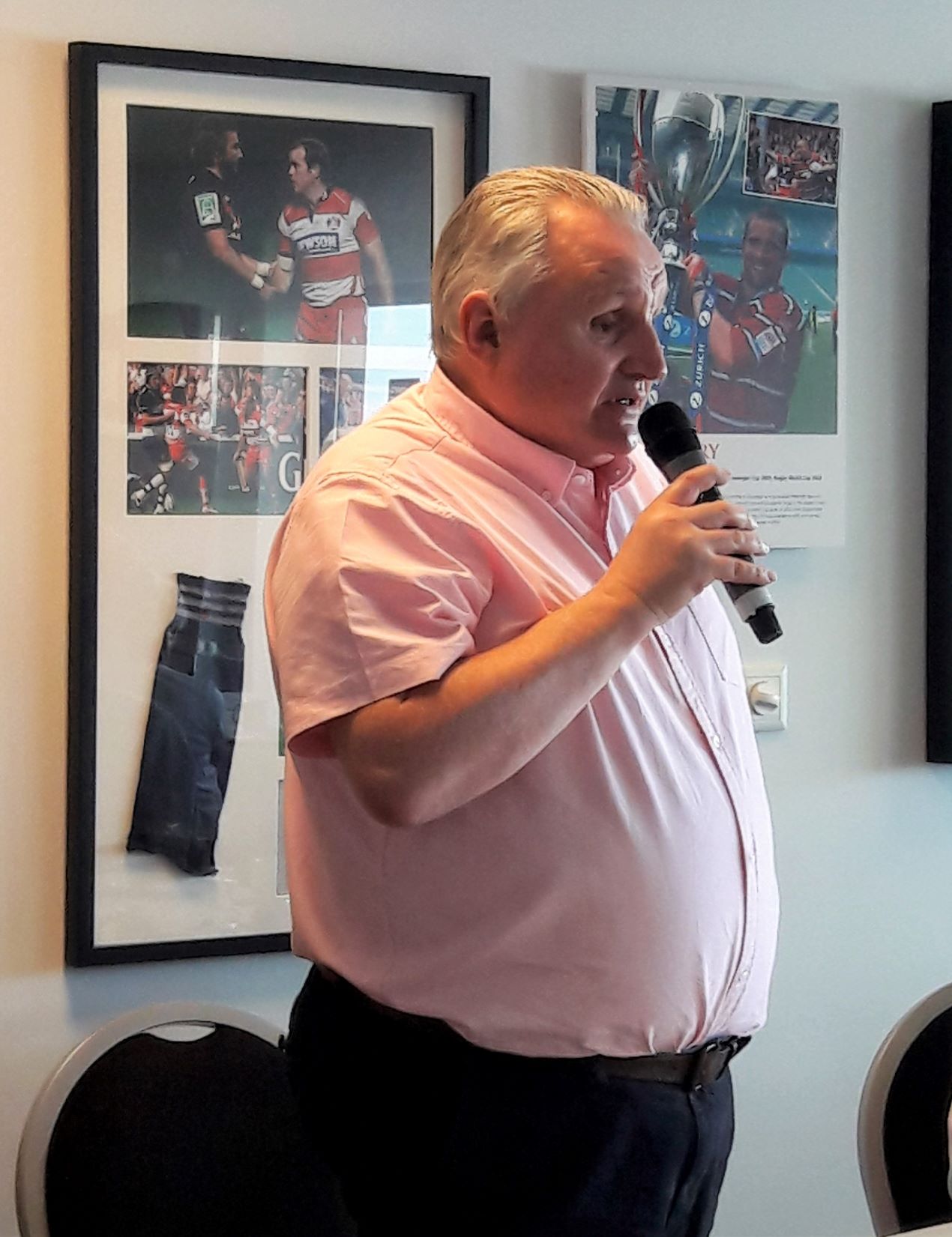 Alun Davies, Engagement manager for Gloucestershire speaking at the Make Transport Accessible forum. He is standing up, talking into a microphone. He is wearing a short sleeve, pale pink shirt and black trousers. Behind him are framed photographs of rugby players on the wall.