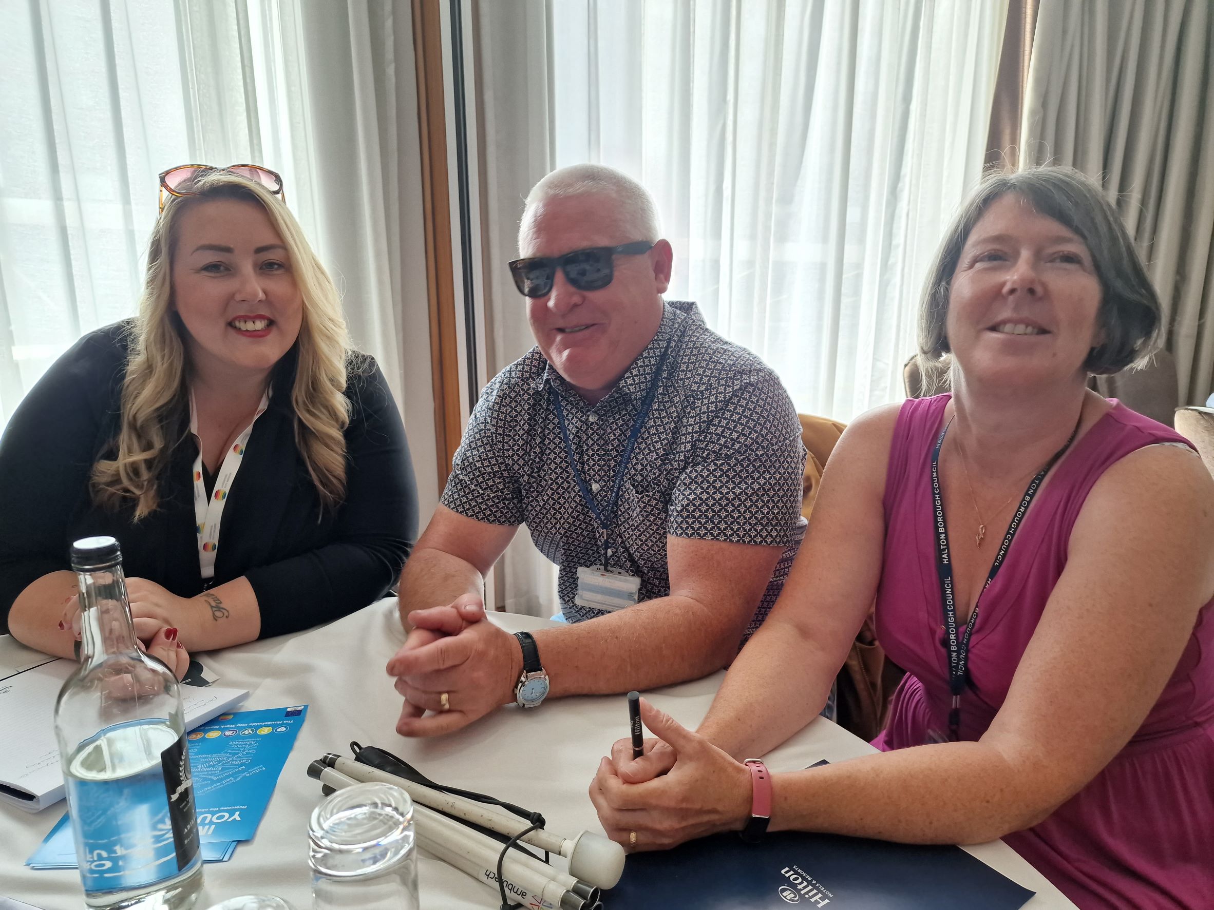 Three delegates sat on a table discussing issues related to blind and partially sighted people. They are looking at the camera and smiling. A folded white cane is on the table.