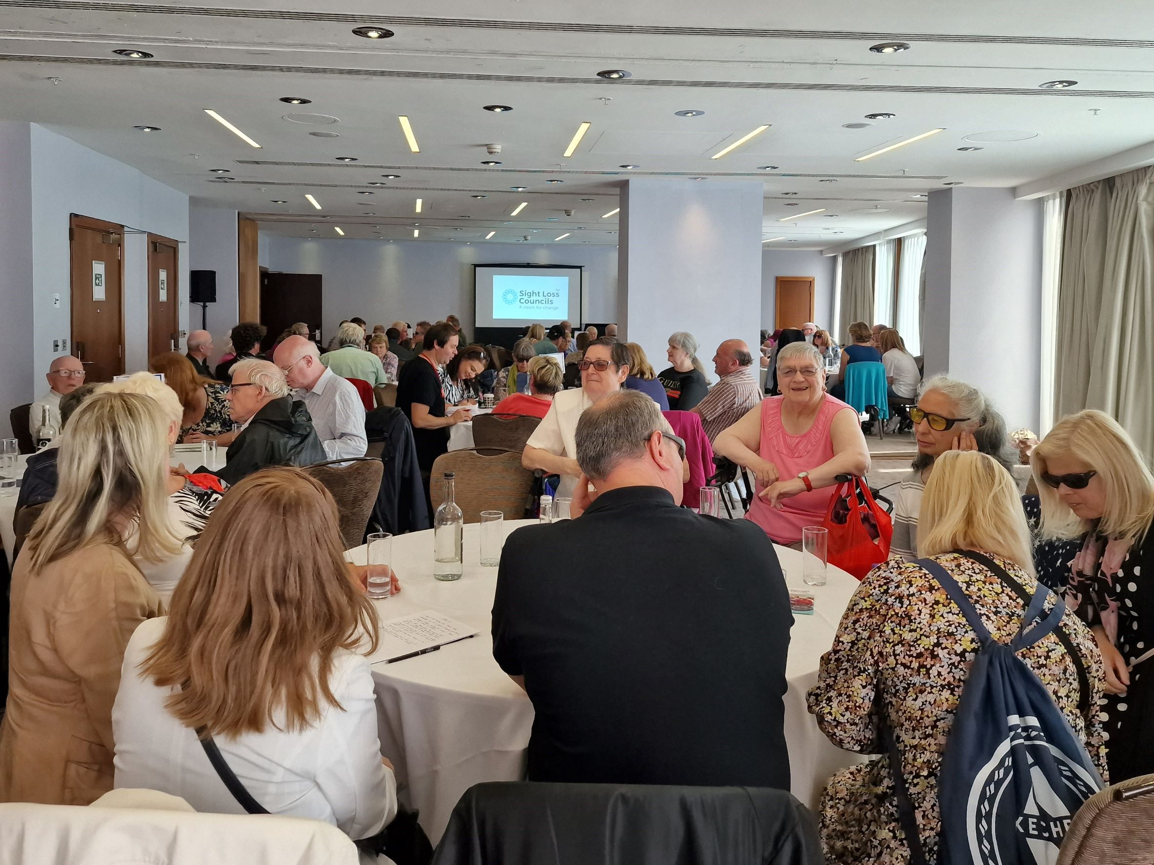 Blind and partially sighted attendees and partners sit around tables discussing key topics focused on employment, transport, health and wellbeing, and accessibility.