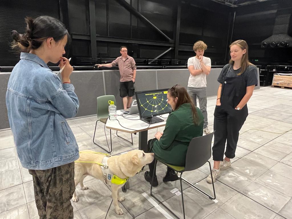 Essex Sight Loss Council member, Samantha Leftwich, is sitting at a desk in front of a computer screen which shows a yellow graph. She is looking down at her guide dof. There are four other attendees and research members standing around her.