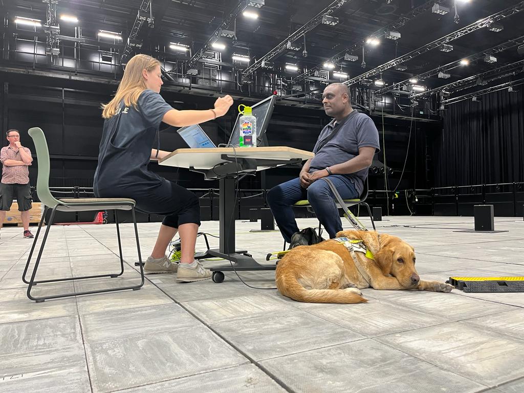 London Sight Loss Council member Haren Thillainathan at UCL Pearl. He is sitting at a desk in the middle of a room, his guide dog is lying on the floor. A member from UCL Pearl is on the other side of the desk, mid conversation. There are two computer screens on the desk.