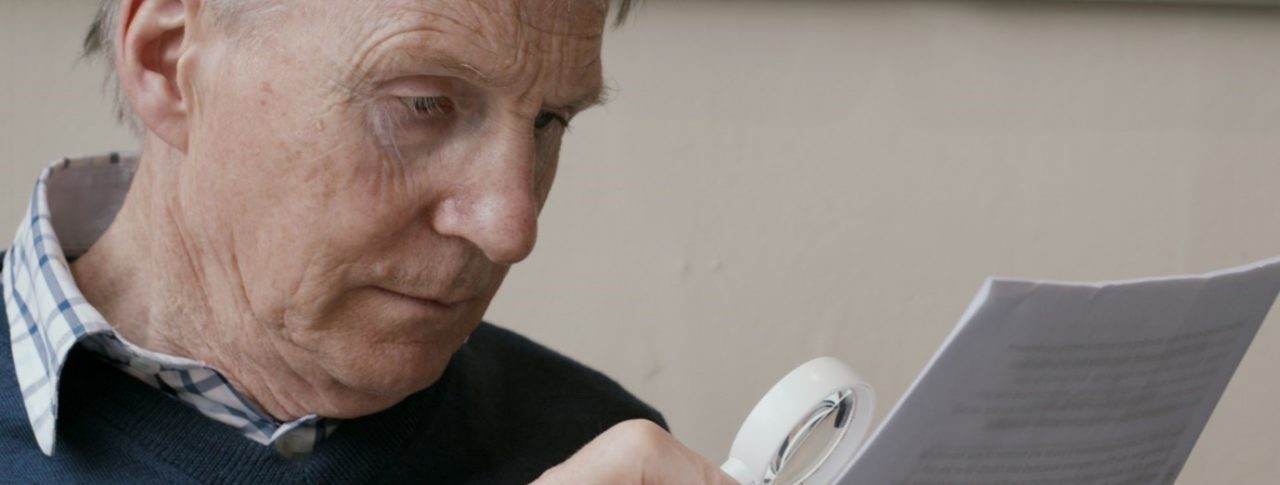 A man in a blue jumper reads from a piece of paper using a magnifier