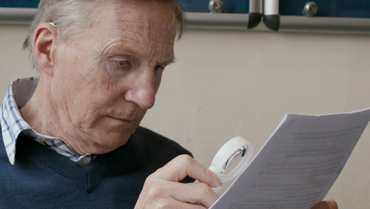 A man in a blue jumper reads from a piece of paper using a magnifier