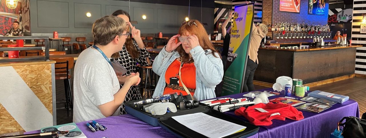 Retailer trying on simulation spectacles, stood with Sight Loss Council members at their stall.