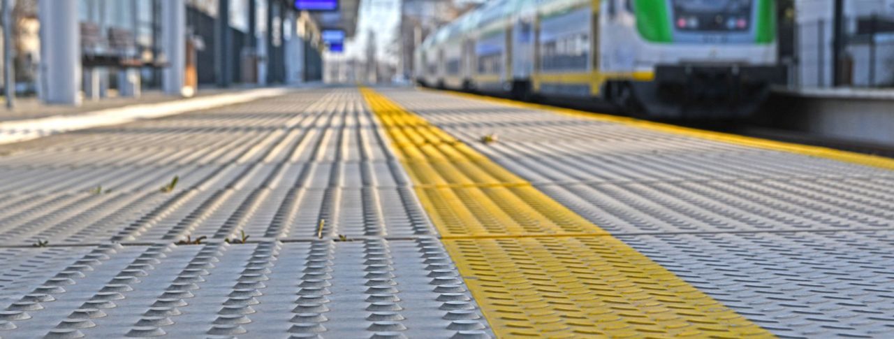 Image shows a train platform with tactile markings. Taken from ground level, it is a close up of the markings running along the platform. In the background you can see the platform screens, a train pulling in and overhead wires.