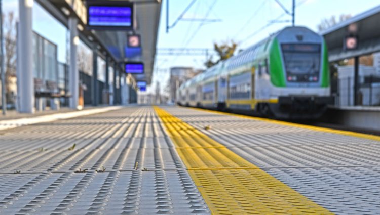 Image shows a train platform with tactile markings. Taken from ground level, it is a close up of the markings running along the platform. In the background you can see the platform screens, a train pulling in and overhead wires.