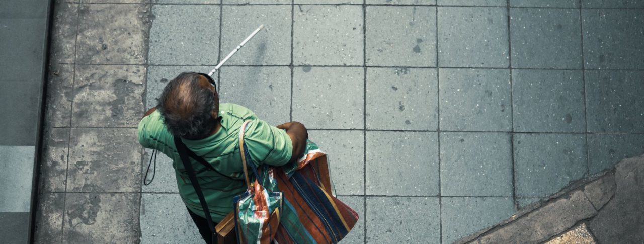 Overhead shot of a blind person walking on a sidewalk. They are using a cane and have a big shopping bag over their shoulder.