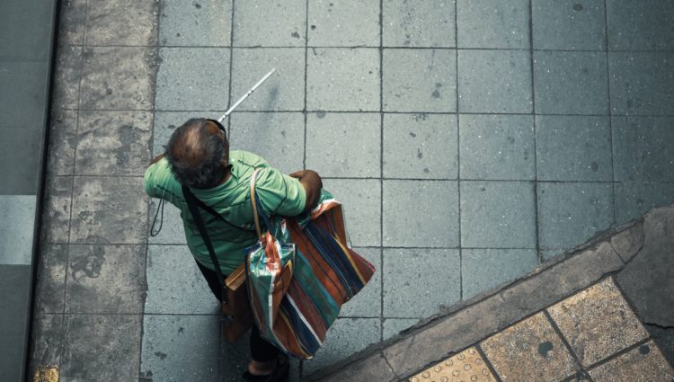 Overhead shot of a blind person walking on a sidewalk. They are using a cane and have a big shopping bag over their shoulder.