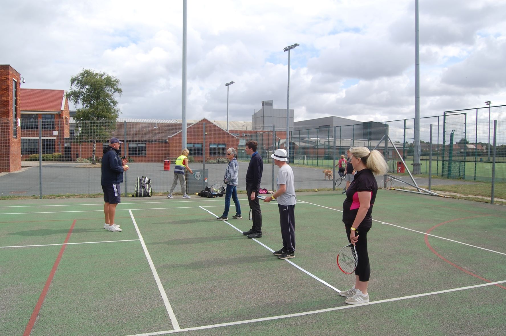 Attendees standing on a tennis court, holding tennis rackets, listening to the coach.