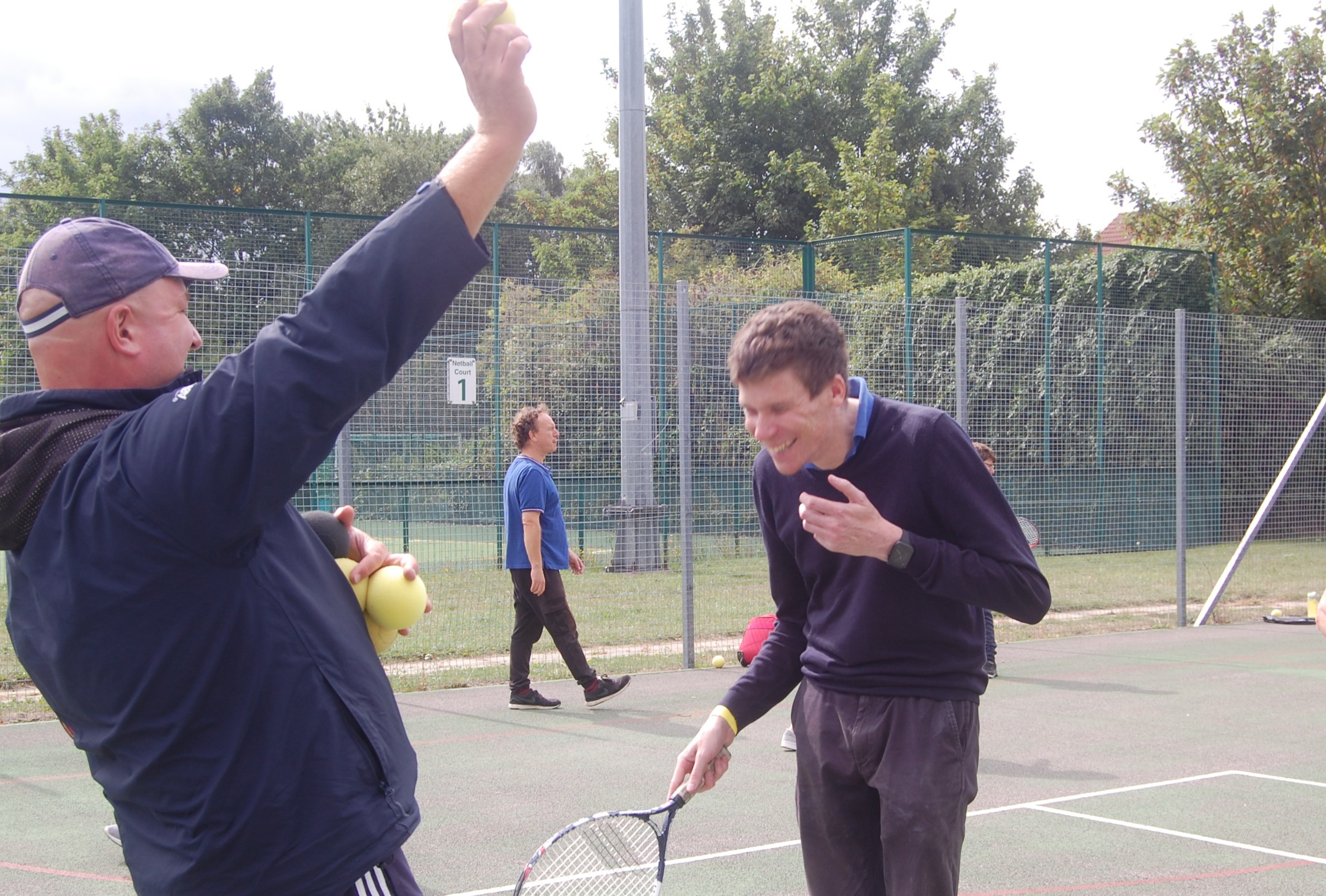 Image shows an attendee standing with a tennis coach, racket in hand. The coach is holding tennis balls in one hand, his other arm raised. The attendee is laughing,