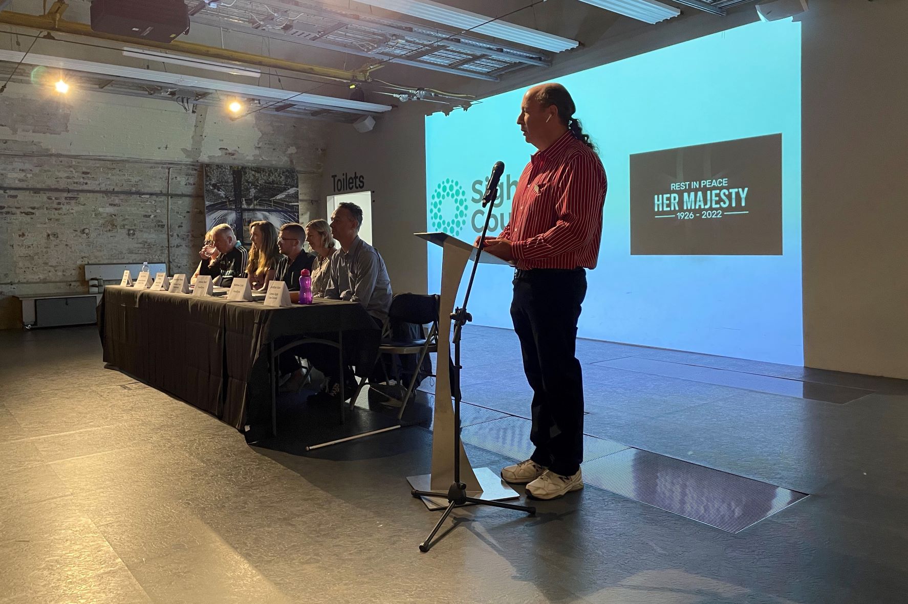 Jonathan Abro, SLC member and event organiser, standing at a plinth, addressing the audience. To the left sit the panel of speakers.