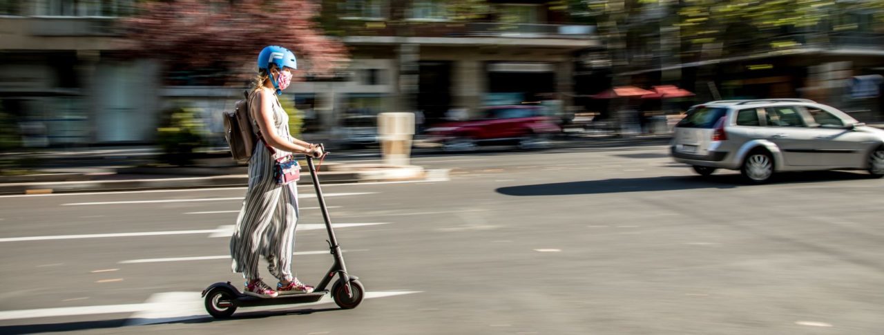 Woman riding an e-scooter down a main road, she is wearing a blue helmet. In the background trees, cars and buildings are blurred out.